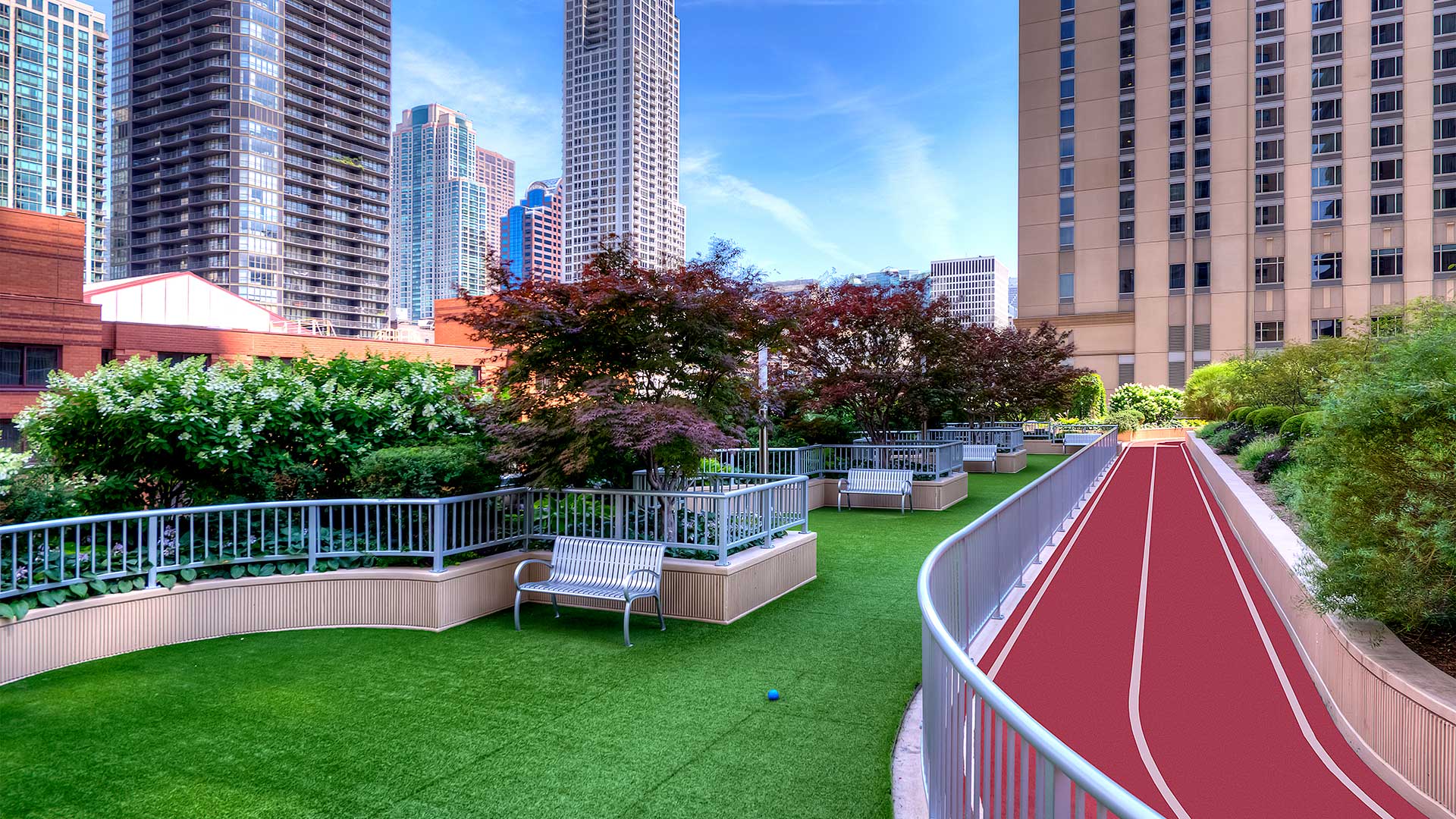 Looking down along the outdoor running track and dog park. The track runs along the right and the green turf of the dog park on the left. City buildings are seen in the background.