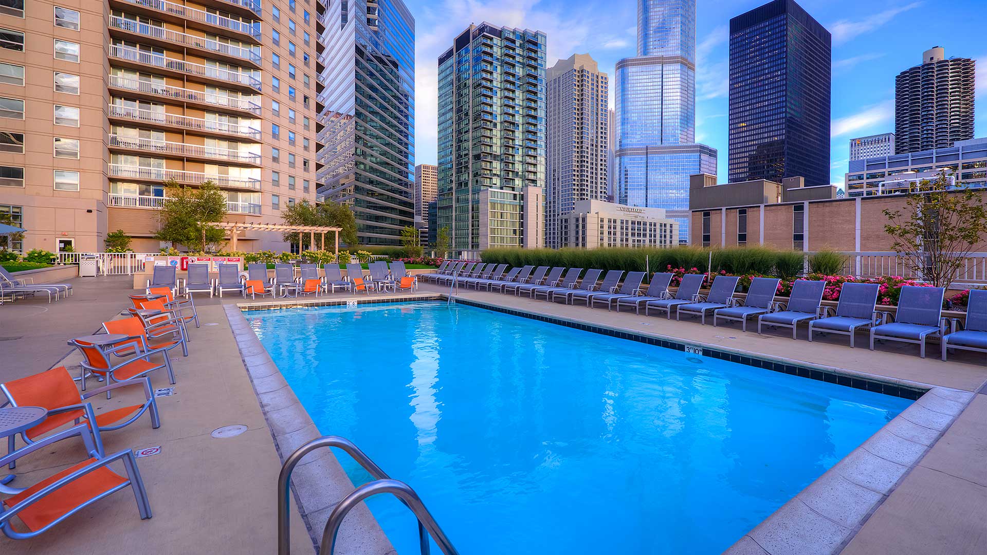 Looking across the outdoor pool late in the day. There are lounge chairs around the pool and the apartment tower rises off to the left. City buildings are seen beyond the deck to the right.