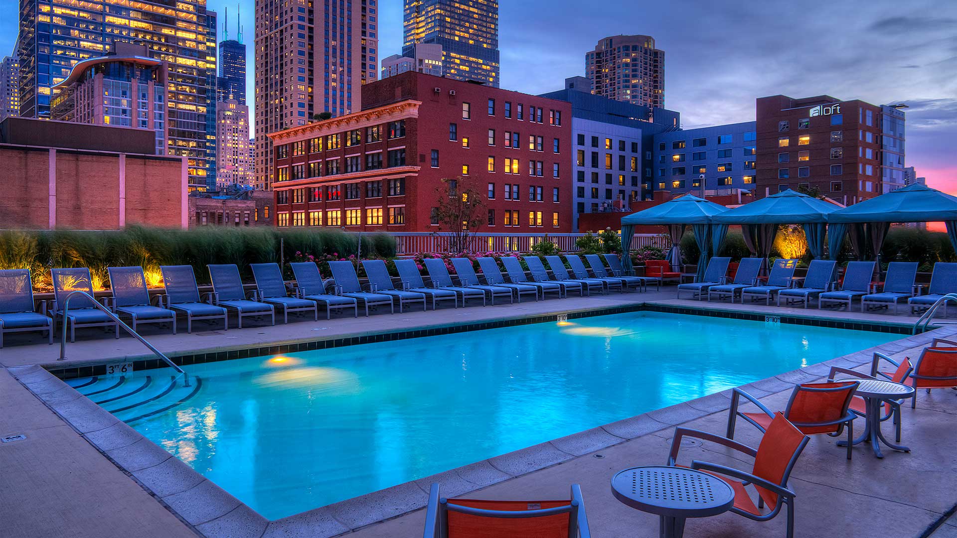 Looking across the outdoor pool at Grand Plaza at night. The pool glows blue and has various lounge chairs around it. City buildings make up the background, lit up at night.