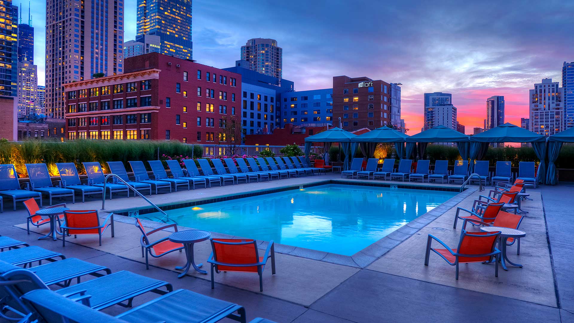 An outdoor pool at dusk. Lounge chairs surround the pool on all four sides. City buildings are seen in the background with lights on.