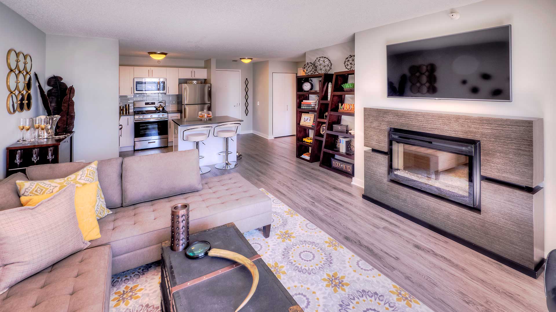 Looking across the living and kitchen in a residence at Grand Plaza. A couch and coffee table are near on the left with a fireplace and television mounted above across. Further back to the left is the kitchen with a bookshelf across from it on the right.