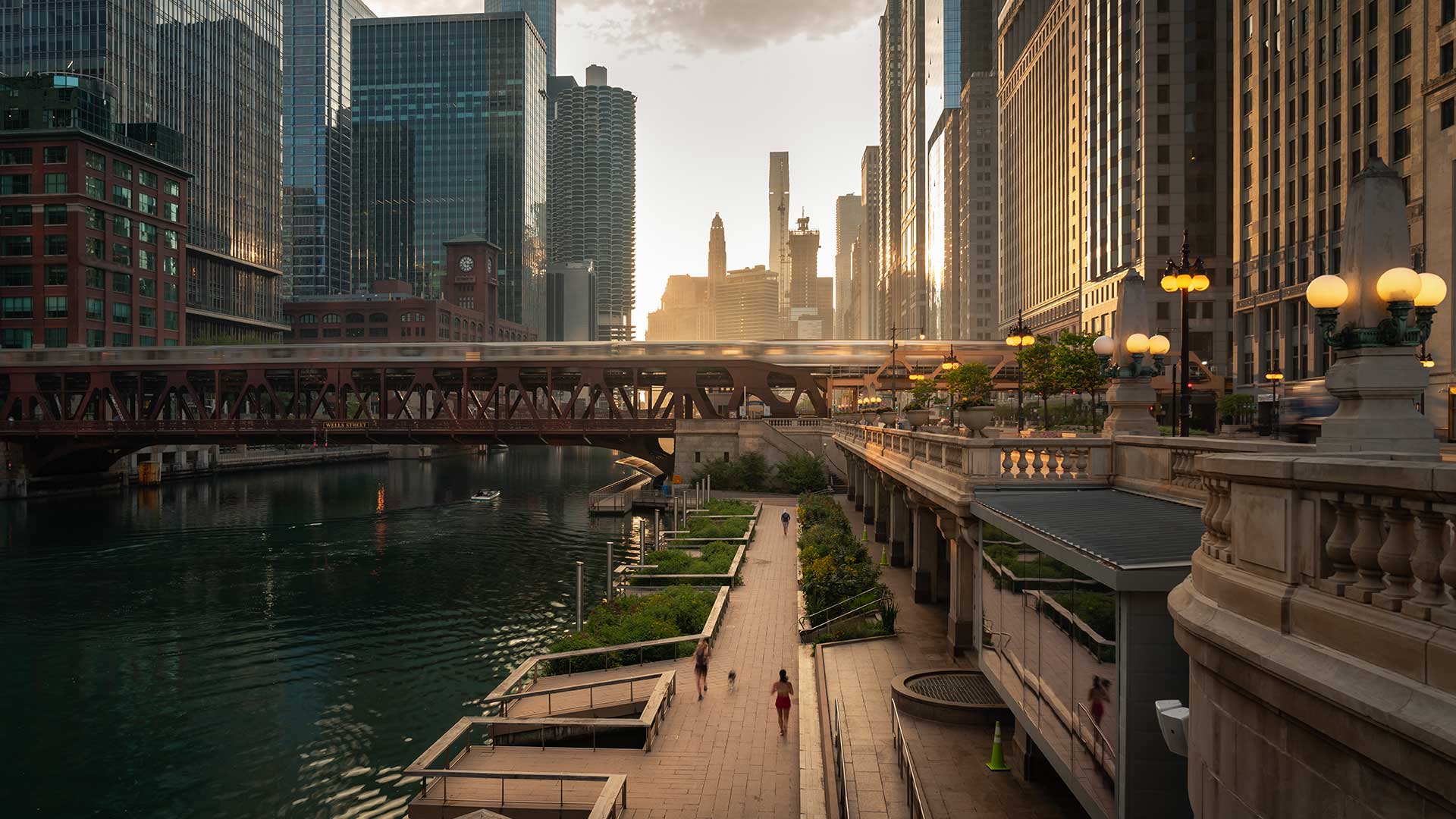 Looking down at the riverfront walkway at dawn in Chicago. There are a few people jogging on the path below. Ahead is a bridge with a train crossing. There are city buildings all around.