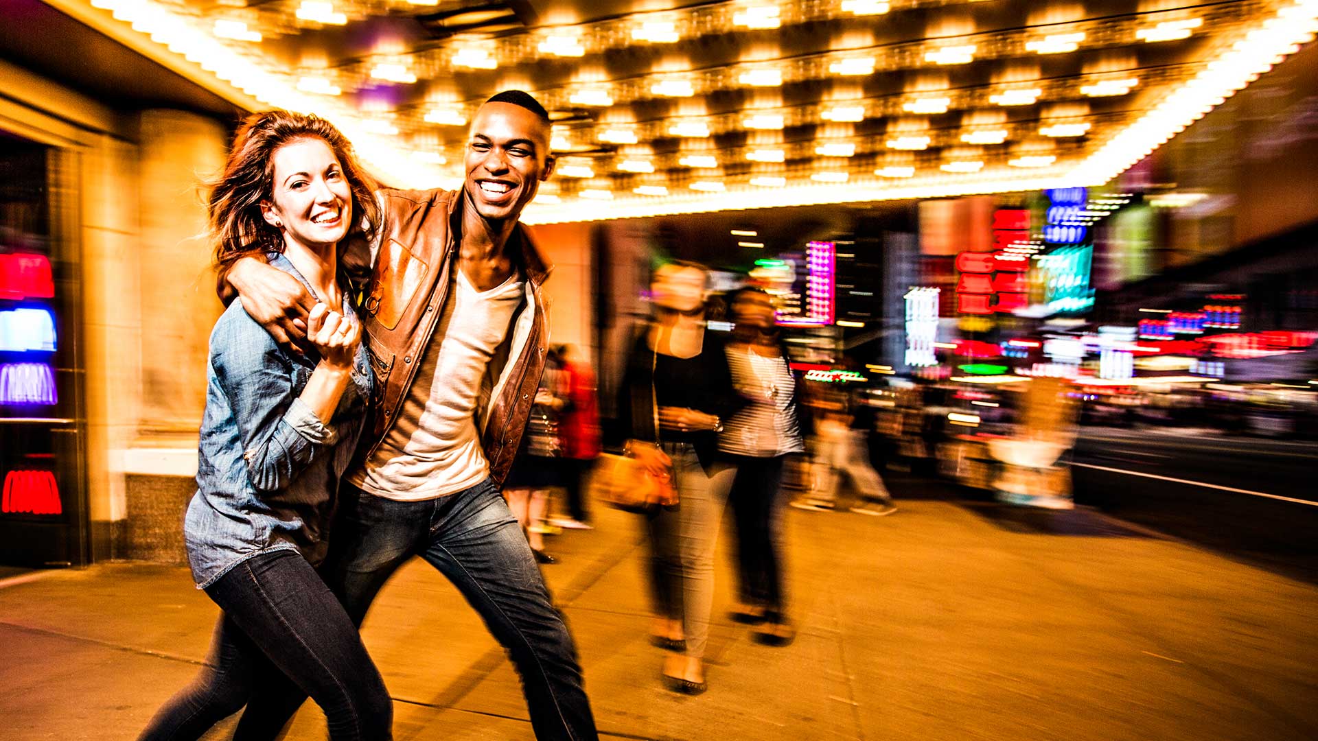 A couple exits a theater and smiles to the camera under the lights of the marquee above. City lights are seen blurry in the background.
