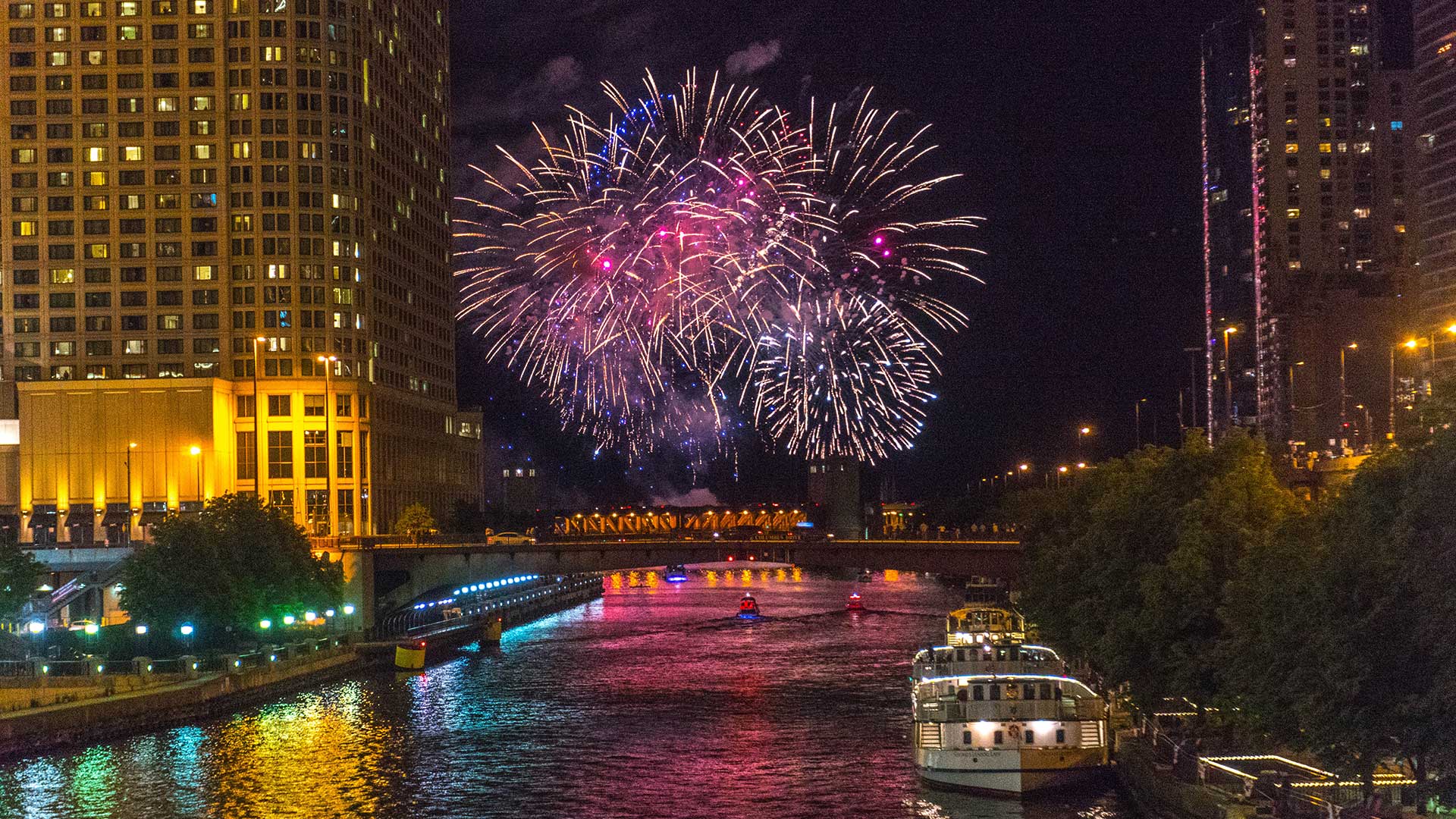 Fireworks light up the night sky over Lake Michigan, as seen from up the Chicago River. There are buildings on both sides of the river and several boats floating.