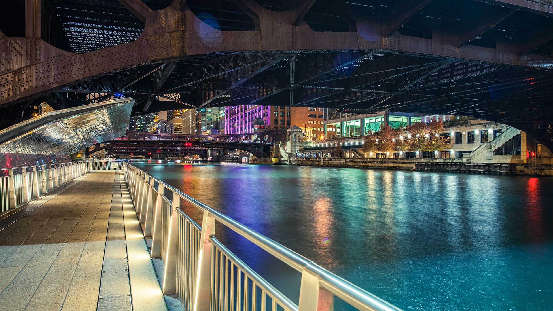 Looking along a pedestrian walkway under a Chicago Bridge at night along the riverfront. The walkway is on the left and the Chicago River on the right. The buildings seen on the other side of river are lit up with many different colors.